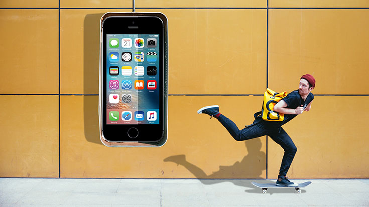 Human skateboarding on a sidewalk wearing a backpack. Behind them is a large depiction of a smartphone secured to the wall.