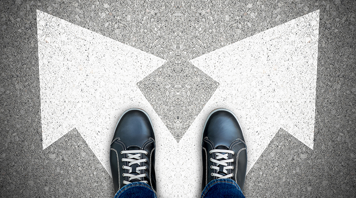 Looking down on two shoes standing at the base of two arrows pointed in different directions.