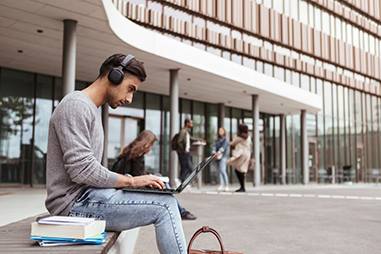 Man sitting outside a building using a laptop and wearing headphones