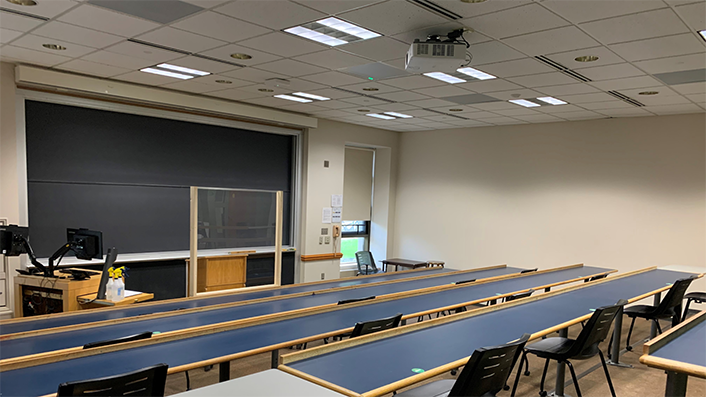 long tables with chairs facing a chalkboard and screen. Two monitors at the front of the classroom.