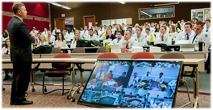 Lecturer in front of classroom and a big monitor showing other rooms of people