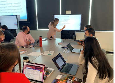 Students around the table with one of them at the large monitor at the end.