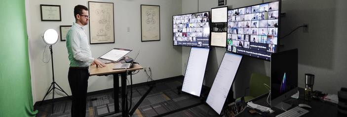 Instructor standing at a lectern facing 2 large monitors. Each monitor is showing a grid of boxes with a student in each.