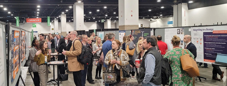 Photograph showing two UTSA students hosting a poster session at the EDUCAUSE Annual Conference. The venue for poster sessions includes many other presenters with their posters, along with crowds of conference attendees talking to presenters about their work