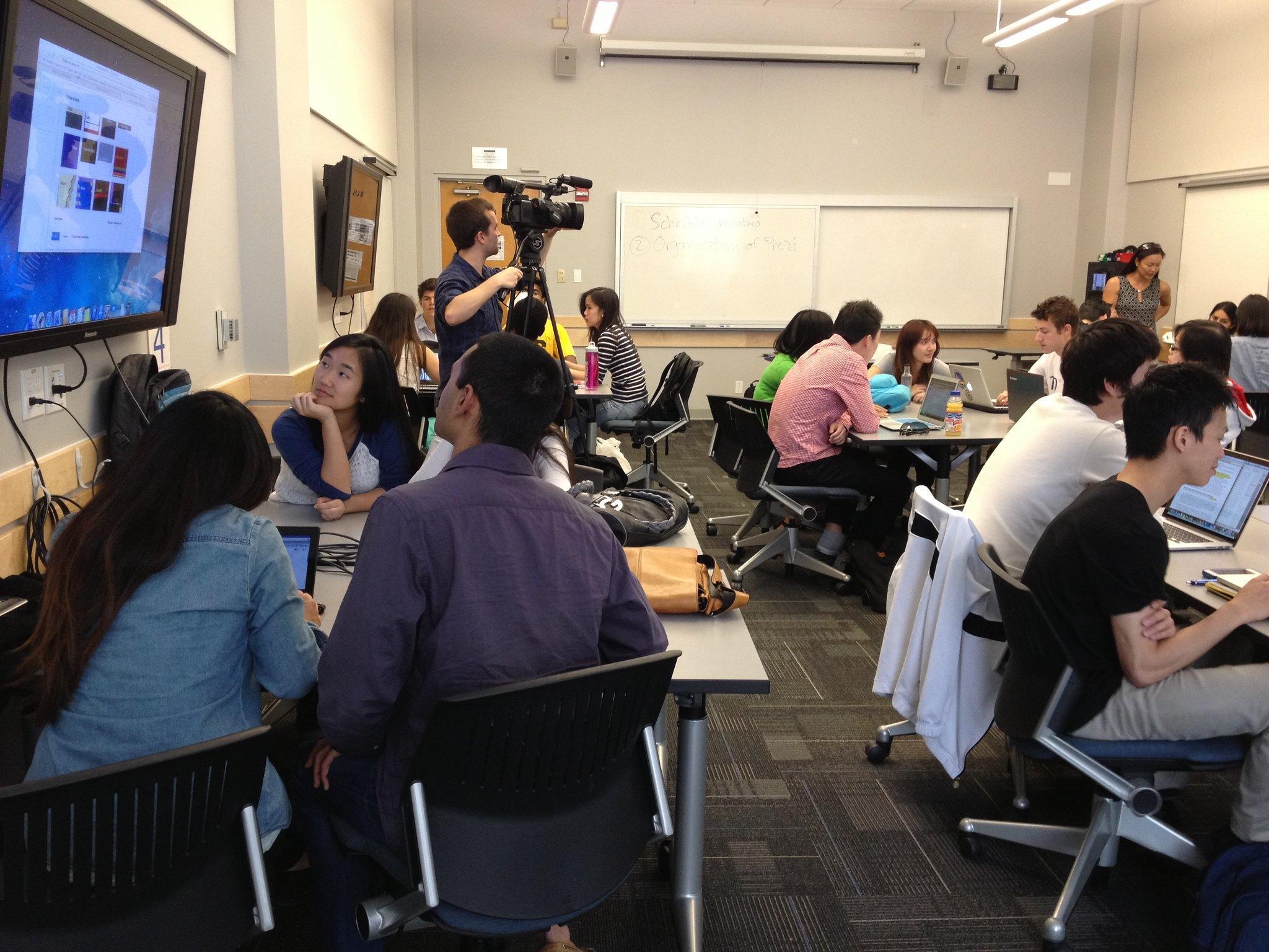Photograph of a classroom with students working at several tables, two large monitors mounted on the wall, and a camera mounted on a tripod.