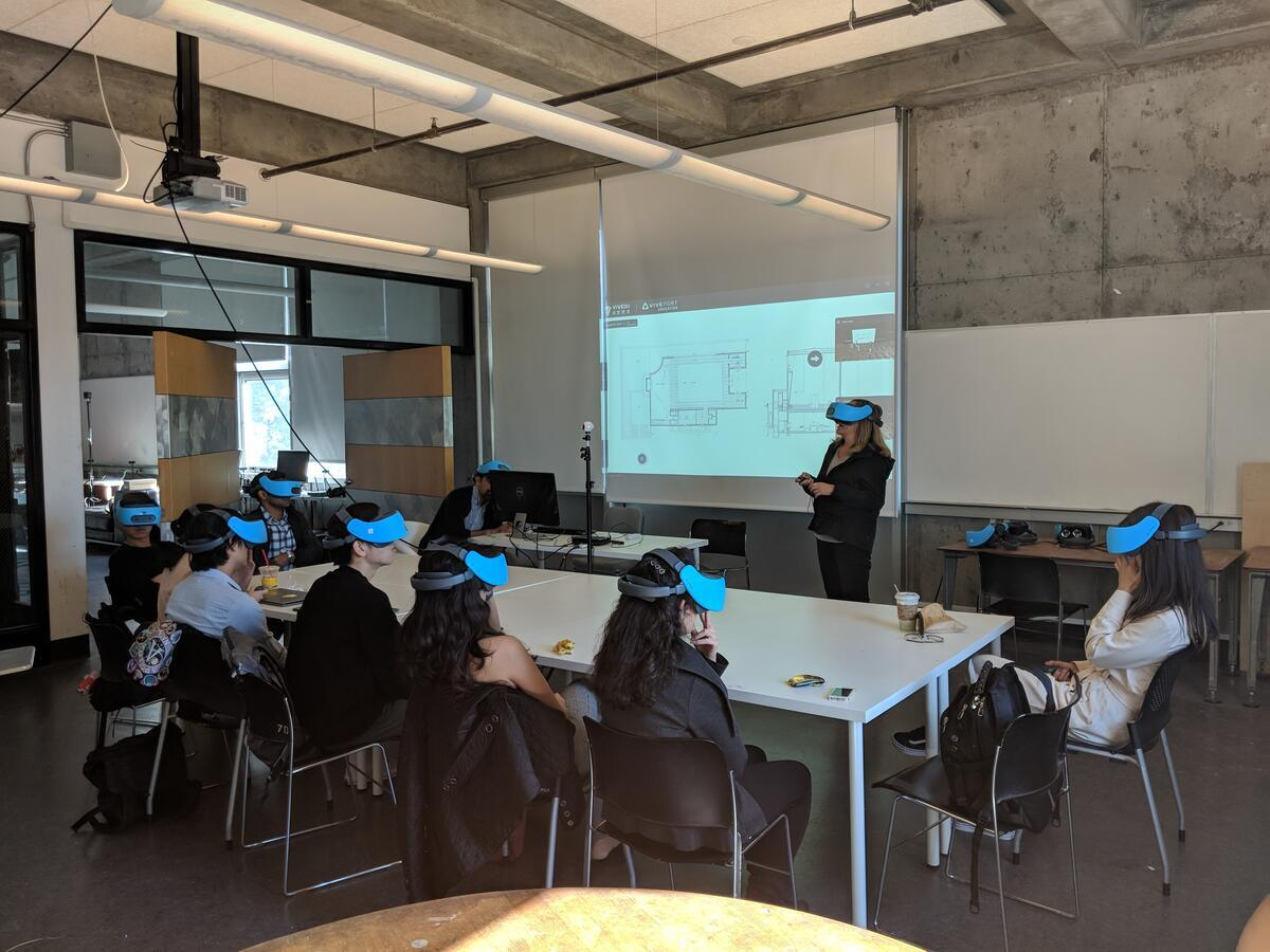 Photograph of an instructor wearing a VR headset standing in front of a small classroom of students, all of whom are also wearing VR headsets.