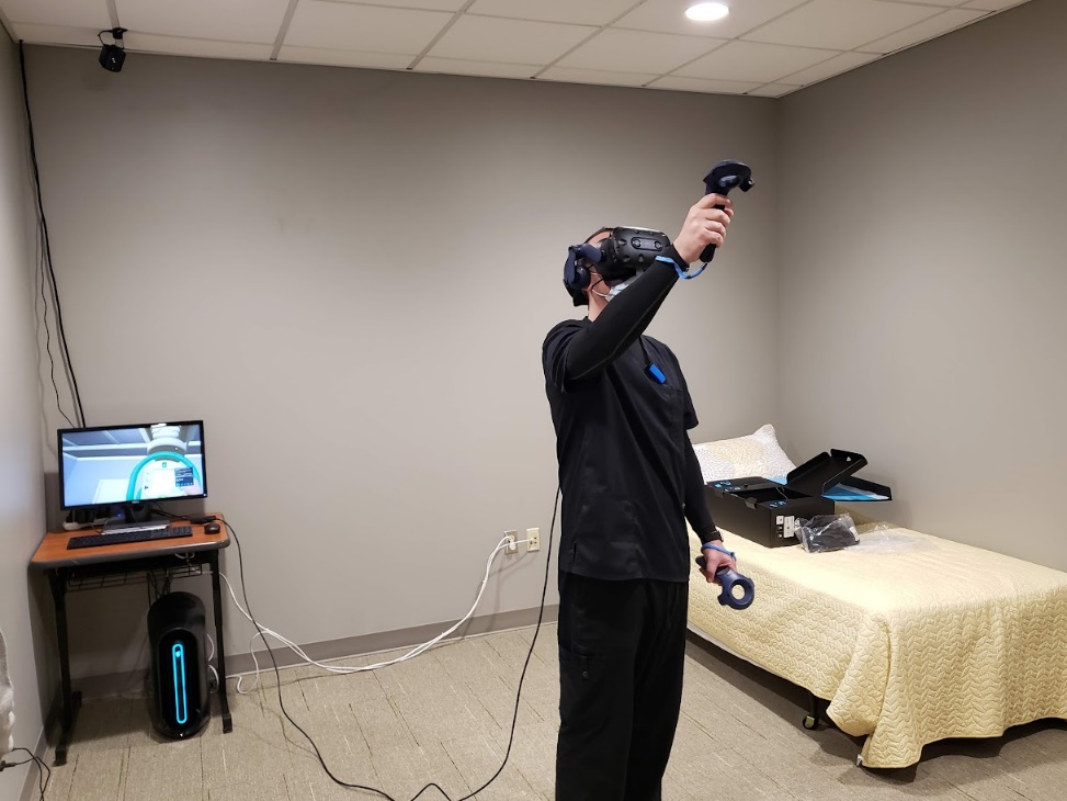 Photograph of a student using the VR equipment. The student wears a VR headset which is connected to a computer in the corner of the small room where the technology is being used. The student is using handheld controllers to manipulate the VR environment.
