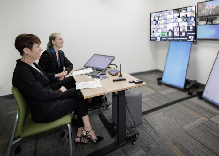 Photograph of the Teaching Space of Tomorrow being used. two people sit at a table, facing several monitors and a camera on the opposite wall that show participants and chat pods. 