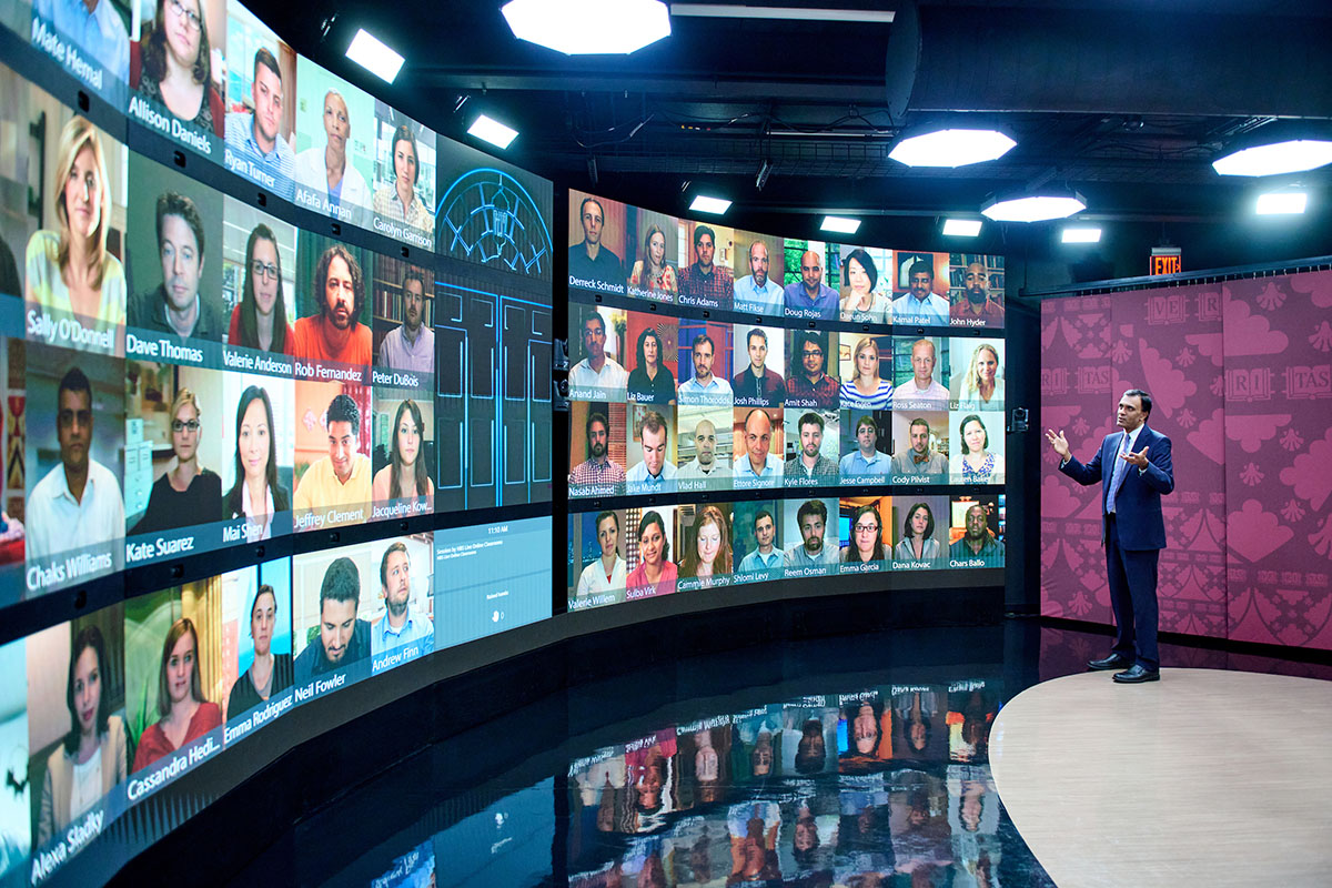 Photograph showing an instructor in an LOC, speaking with students who are shown on a large, curved wall of video screens. 