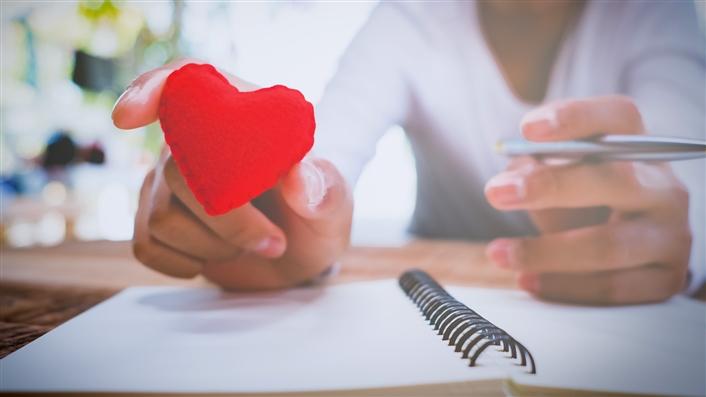photo of person leaning over a notepad and holding a red cutout heart between her thumb and forefinger