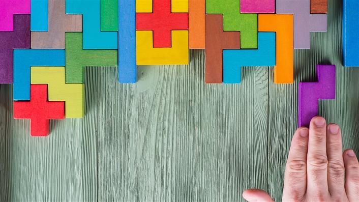 Wood blocks of varying shapes on a board. They are fitted together across the top of the board. A hand is pushing another one into an empty spot which is the right shape.