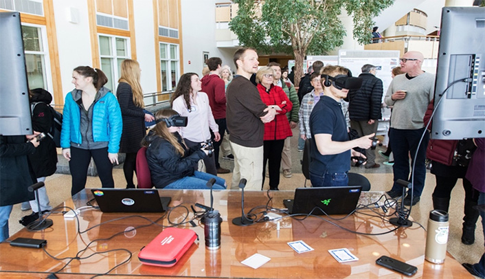 group of people looking at displays.  two people near the front (one sitting and one standing) are wearing VR goggles an have controls in their hands.