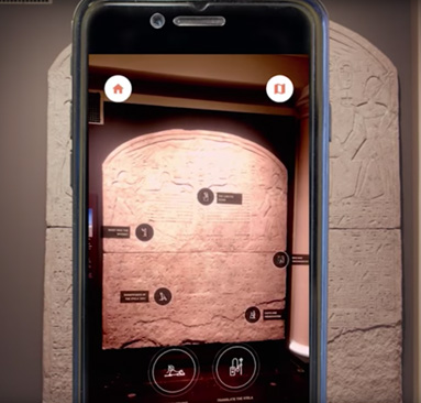 Smartphone being held in front of an artifact. The screen shows the artifact with multiple labels on it (labels not readable in image)