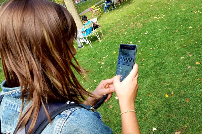 student holding up a smartphone while standing on a large grassy area. other students are seated around the grassy area.