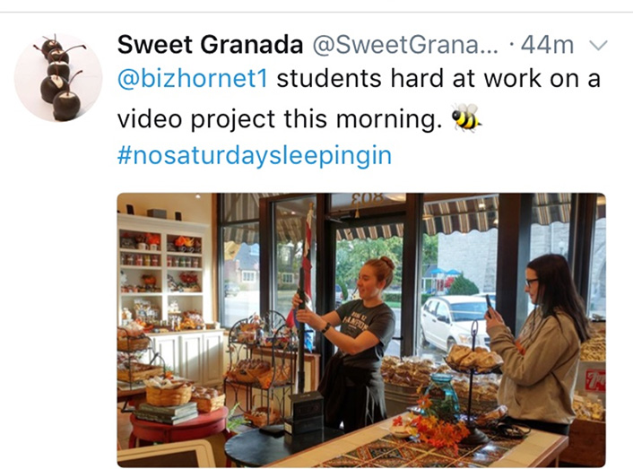 Tweet from Sweet Granada: @bizhornet1 students hard at work on a video project this morning [bee icon] #nosaturdaysleepingin.  An image attached to the tweet shows two students setting up  in a store.