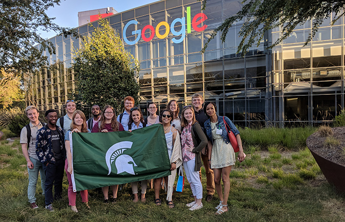 Group of students holding Spartan flag with glass Google building behind them. 