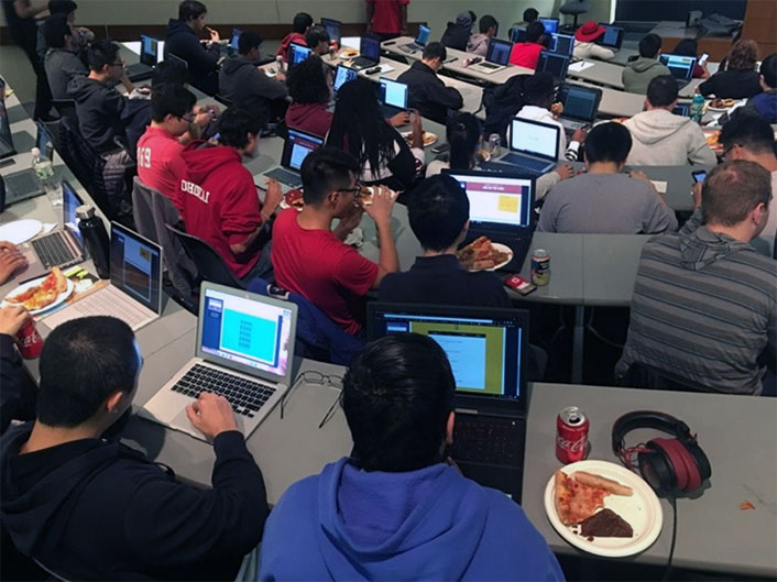 View of classroom with students at all the desks. They each have a laptop open in front of them and a plate of pizza.