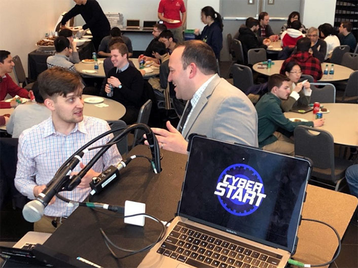 Students at round tables eating. A podium at the front of the room has a laptop open with the CyberStart logo on the screen.