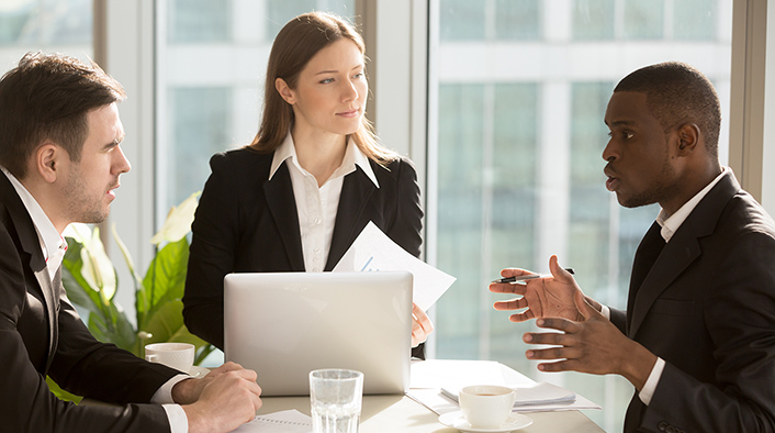 photo of three people in black business suits and white shirts having a conversation at a table