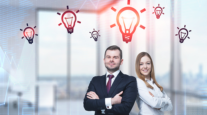 Young businessman and businesswoman standing in blurred office with light bulb sketches and graphs above them.