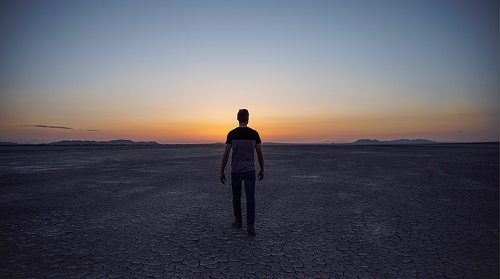 man walking into a sunset across a baked, barren landscape with mountains on the distant horizon