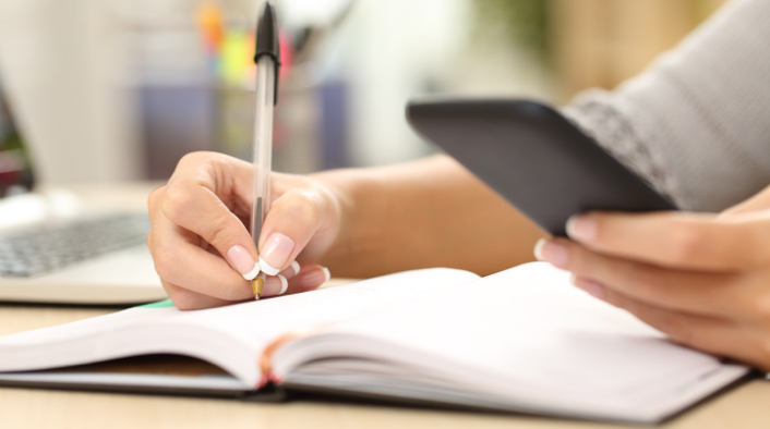 person writing in a book with a pen in right hand while holding mobile device in the left hand