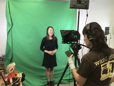 Woman standing in front of a green while someone films her with a video camera.